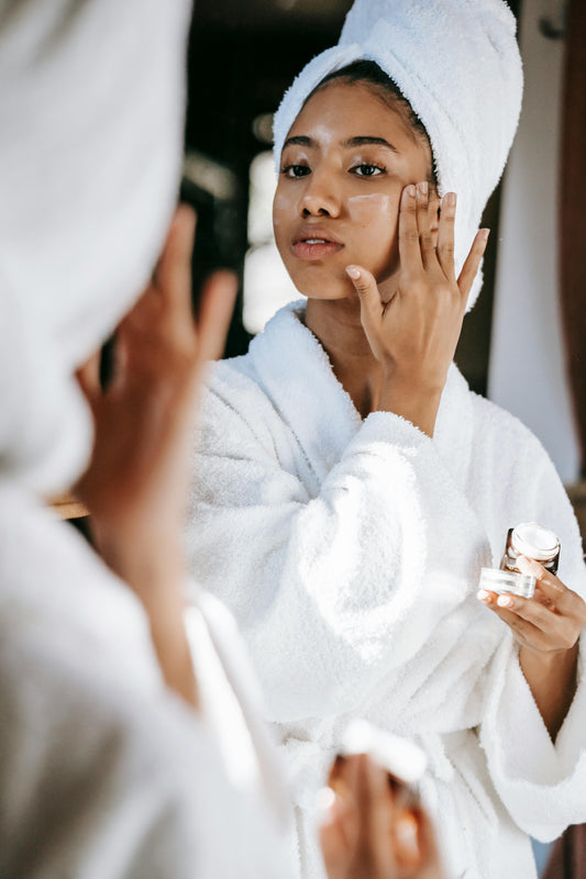 Mujer joven aplicando crema facial frente al espejo con bata blanca